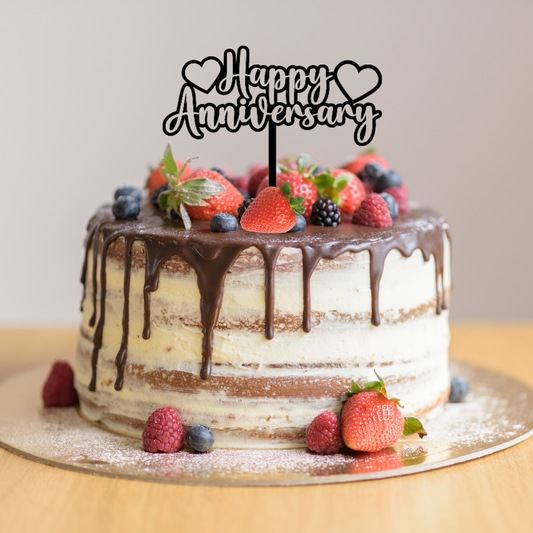 Cake topper with the words: Happy Anniversary and a love heart on either side in black on a cake. Cake topper can be made in different colours.